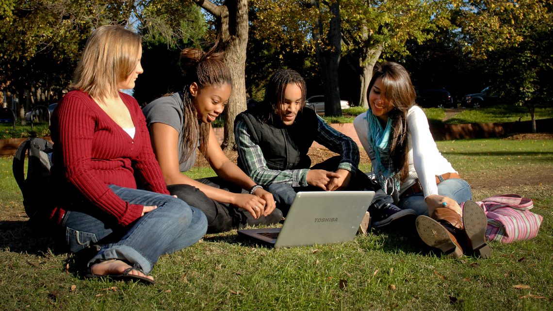 A group of student's sitting outside together as they watch something on a laptop.
