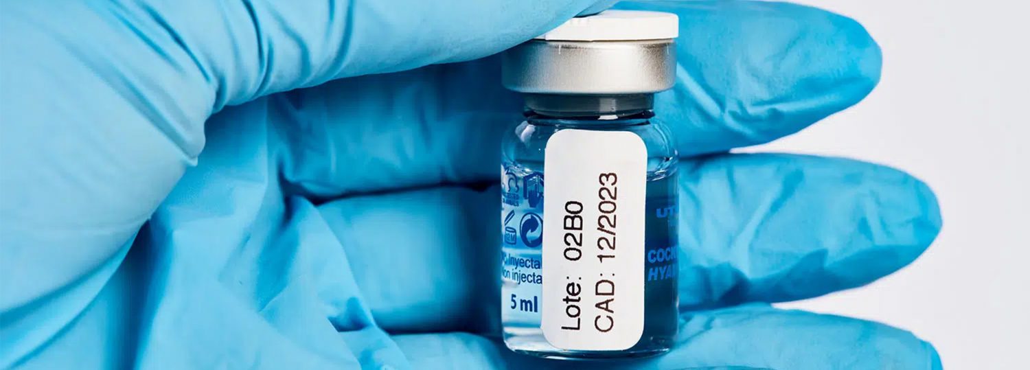 A blue medical gloved hand holding a vial of COVID-19 vaccine