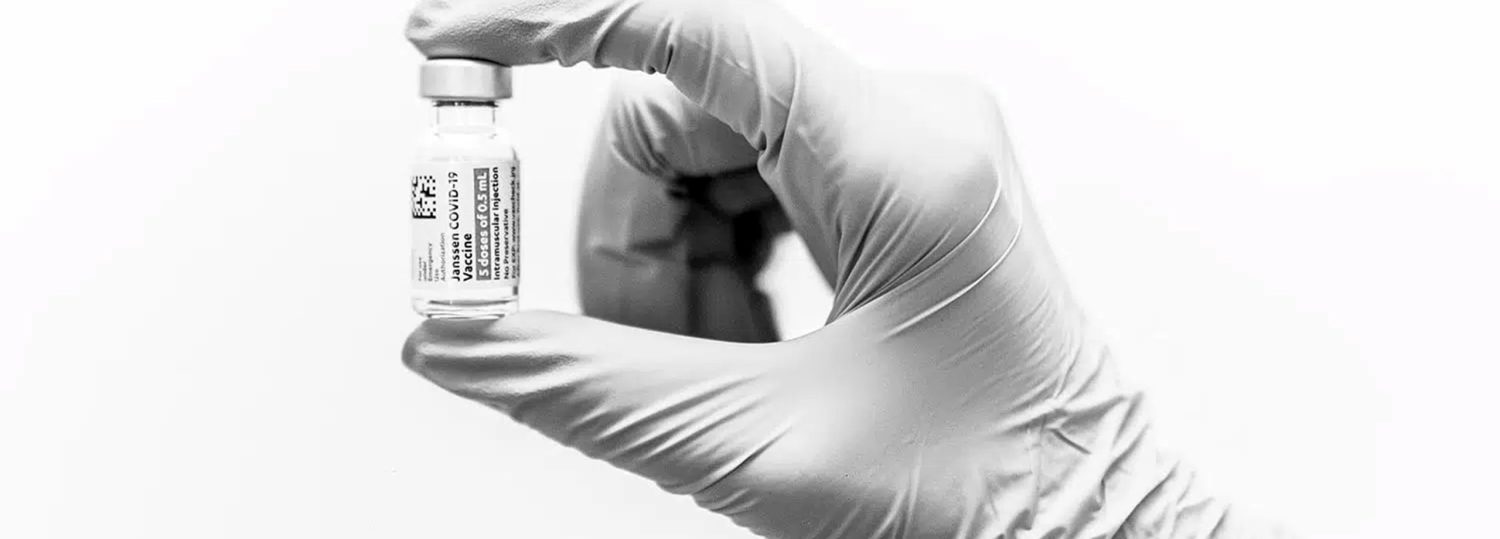 A hand wearing a latex glove is holding a vaccine vial between its index finger and thumb