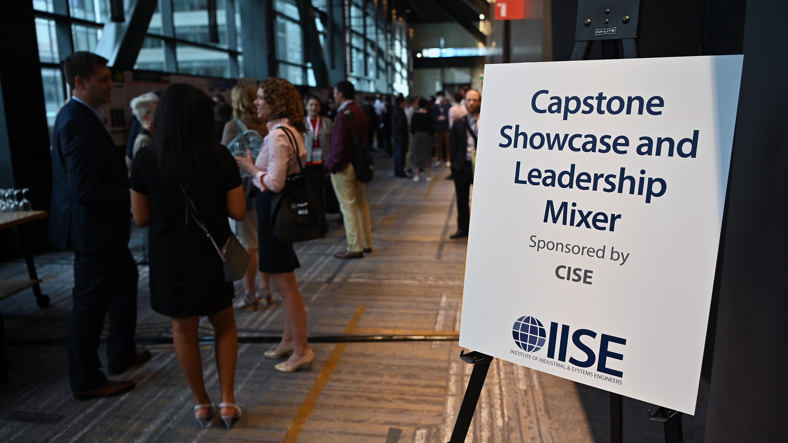 A sign reading, "Capstone Showcase and Leadership Mixer" in front of a long hallway of research posters and people