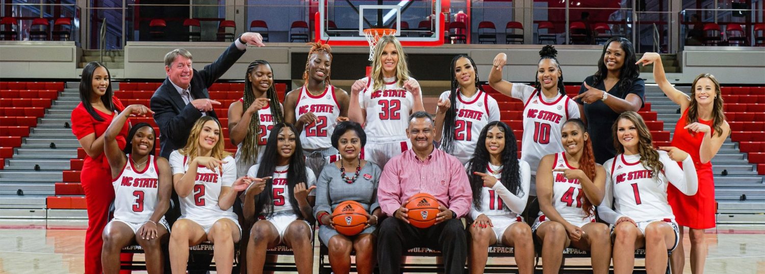 Gayle and Dwain Lanier sitting in chairs on the basketball court surrounded by the NC State Women's Basketball Team and coaching staff who are all pointing at the Laniers