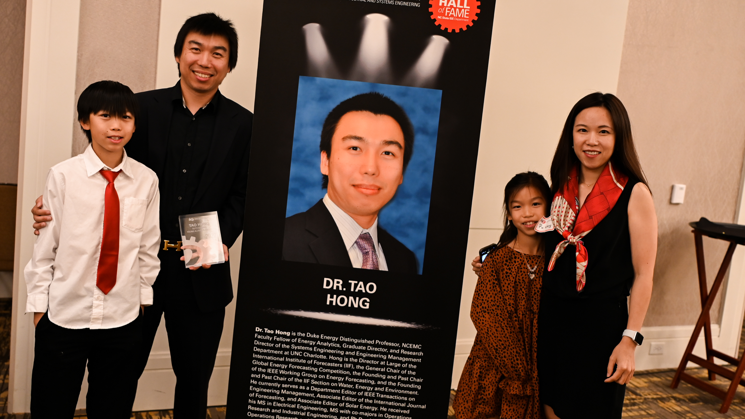 Dr. Tao Hong and his family posing in front of his Outstanding Young Alumni banner