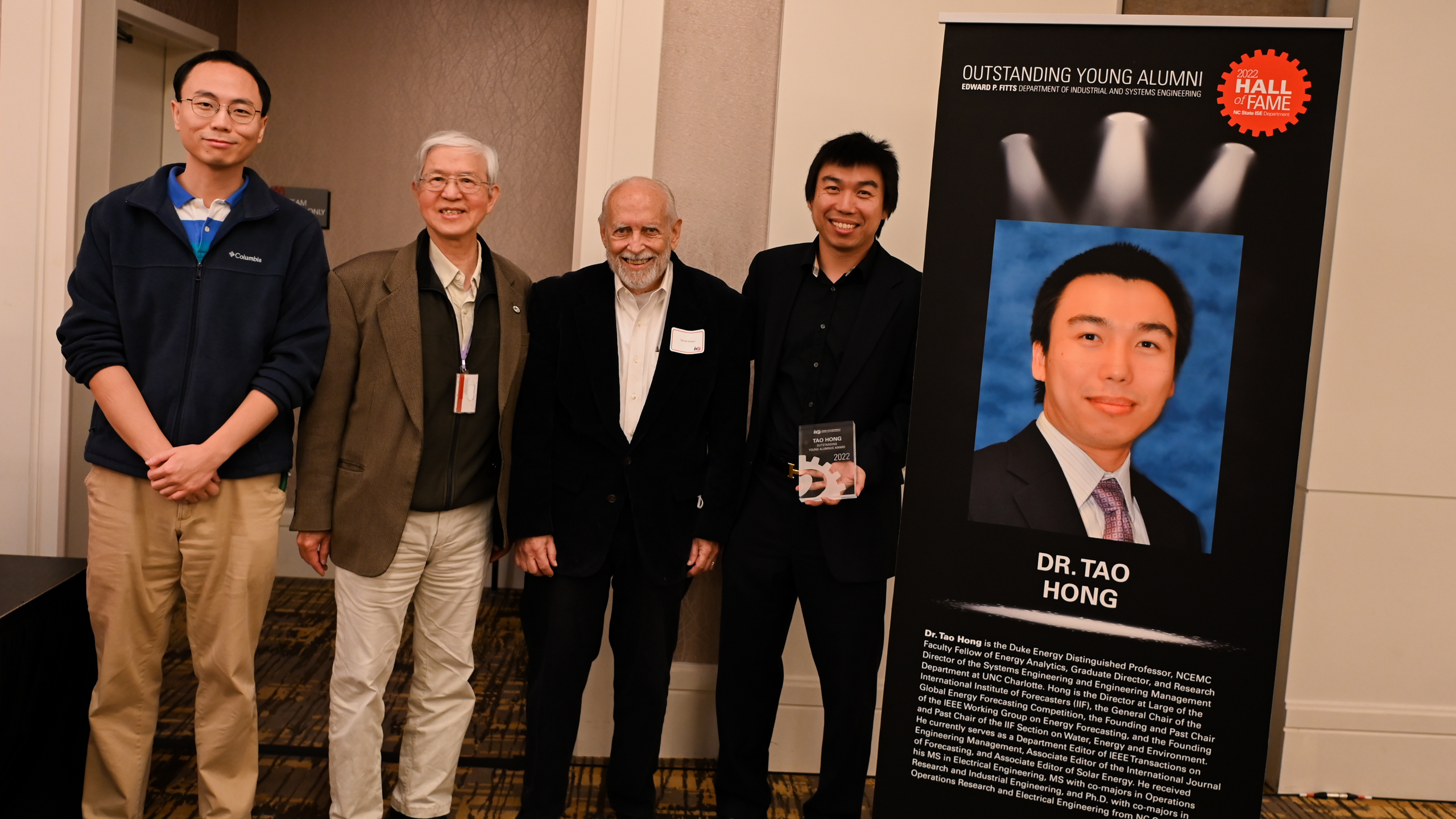 Dr. Tao Hong and faculty members posing in front of his Outstanding Young Alumni banner