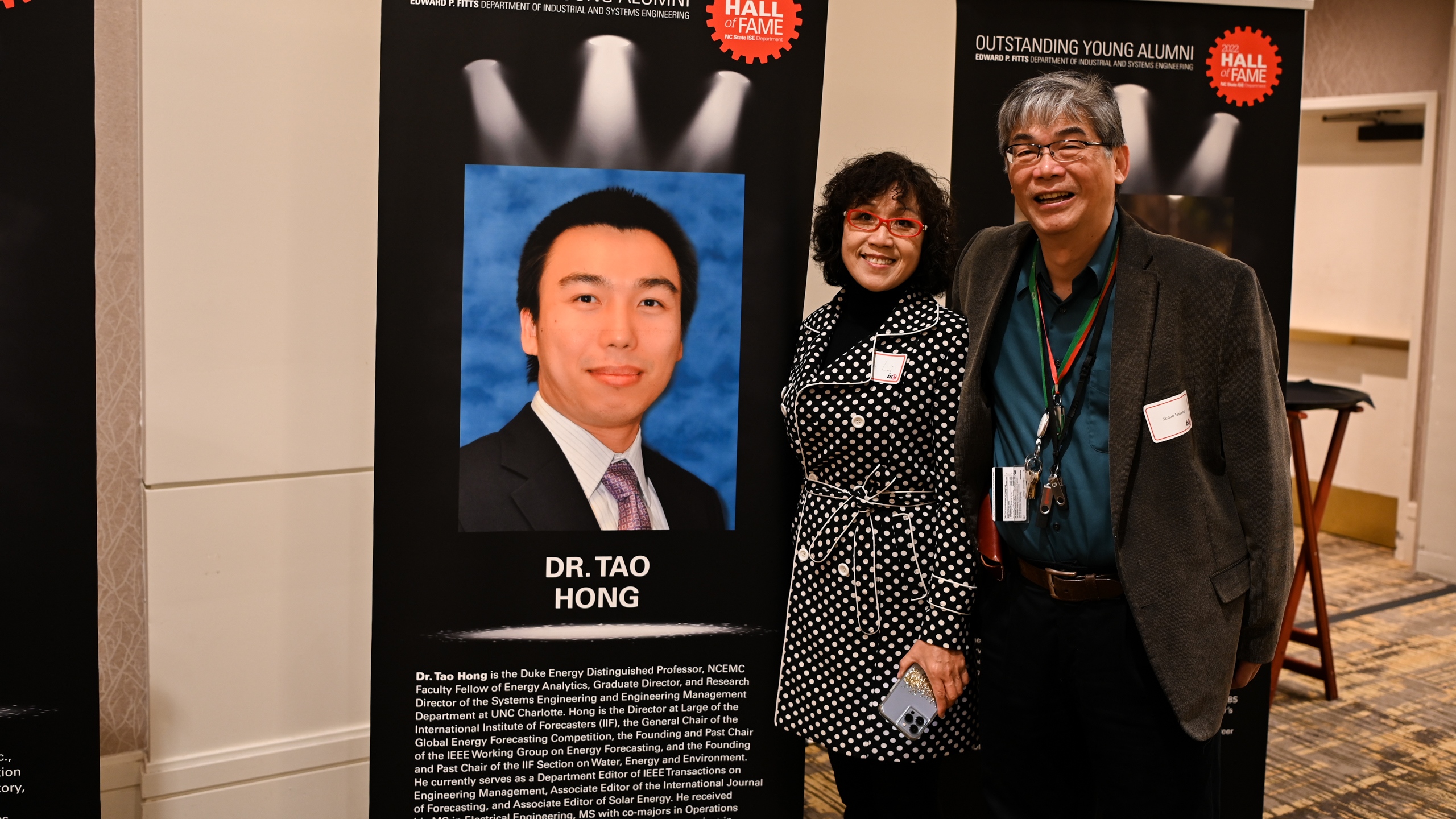Dr. Simon Hsiang and his wife posing in front of Dr. Tao Hong's Outstanding Young Alumni banner