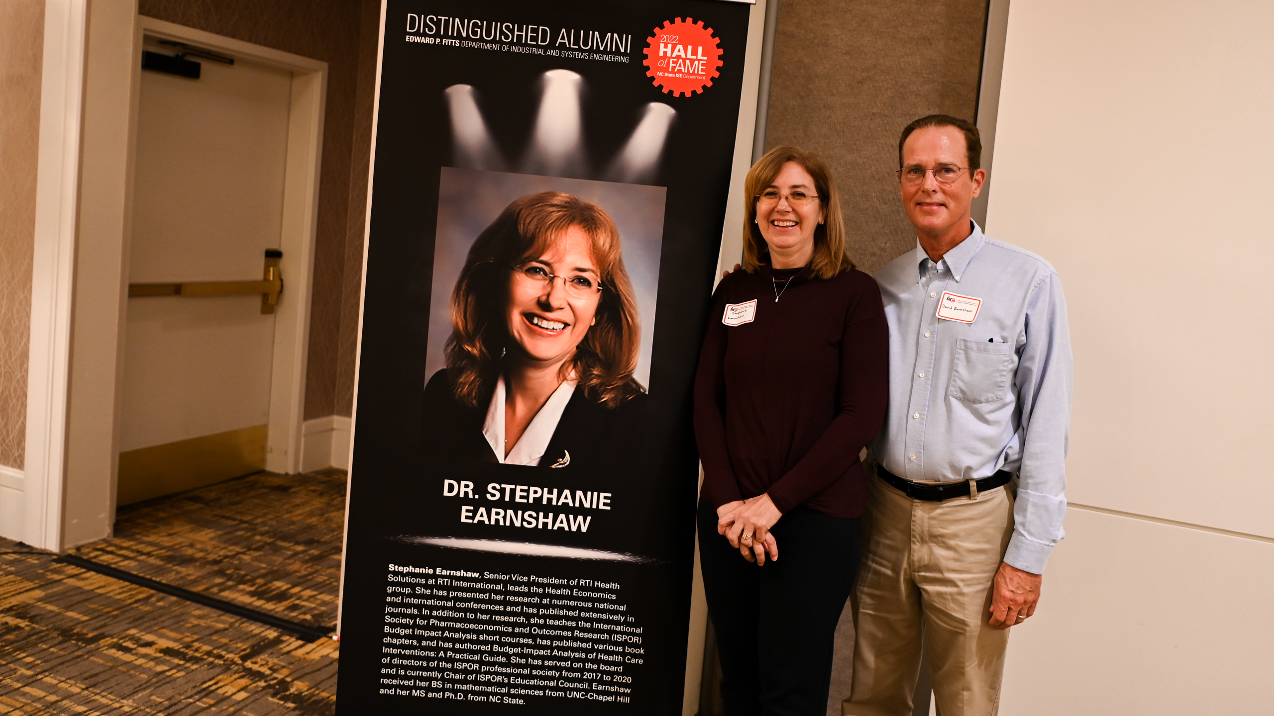 Dr. Stephanie Earnshaw and her husband posing in front of her Distinguished Alumni banner