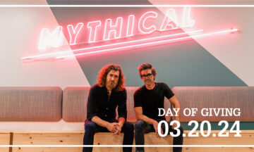 Rhett & Link sitting on a couch under the giant neon light that spells "Mythical."